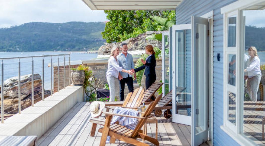 Real estate agent showing a mature couple a new house. The house is contemporary. All are happy and smiling and shaking hands. The couple are casually dressed and the agent is in a suit. Waterfront can be seen in the background